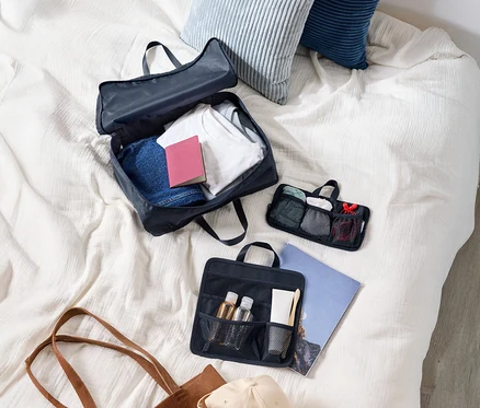 Draufsicht auf einen Koffer mit Kleidung, zwei Kissen, eine Kosmetiktasche, eine Tragetasche und eine Kappe auf einem Bett.