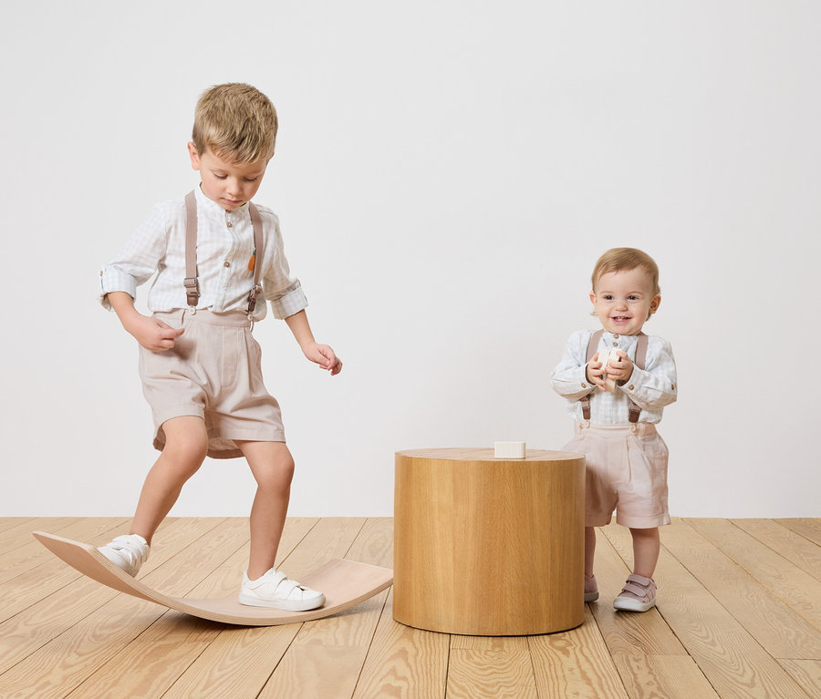 Zwei Kinder, ein Junge balanciert auf einem Holzbrett, ein Baby steht neben einem Holztisch und hält einen Spielzeugblock.