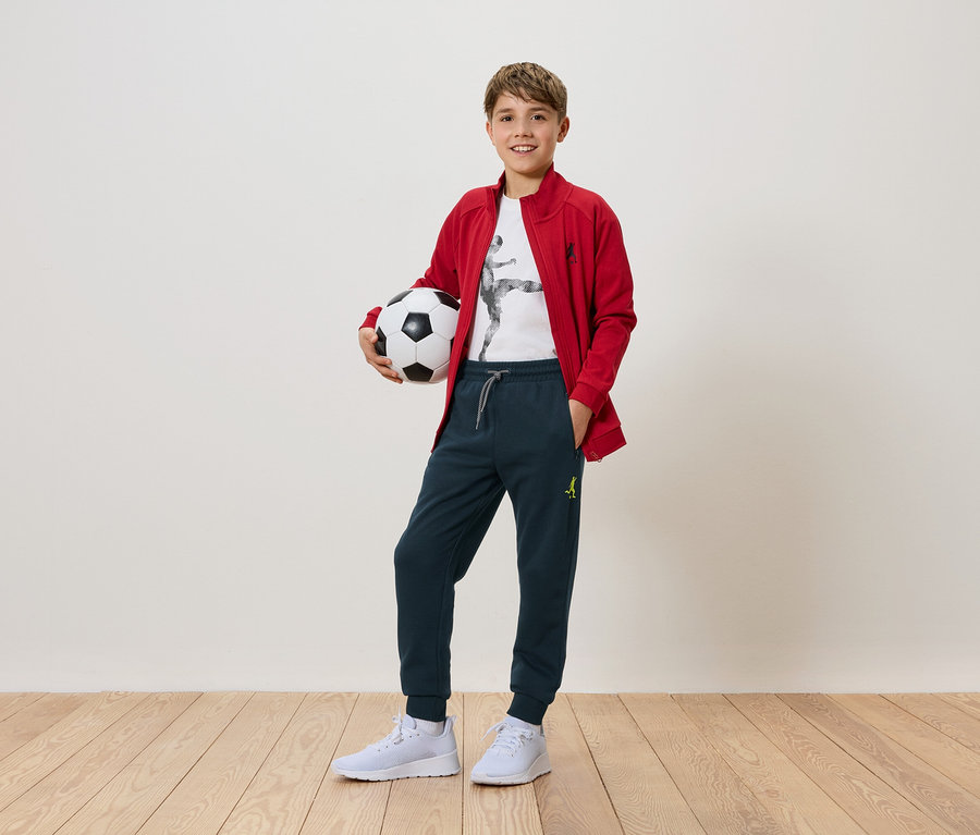 Ein Junge hält einen Fußball in den Händen. Er trägt eine rote Jacke, eine Hose und weiße Turnschuhe.