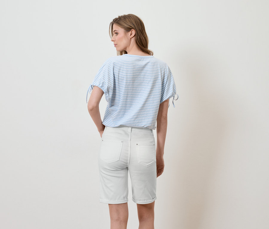 Eine Frau steht mit dem Rücken zur Kamera, trägt ein blau-weiß gestreiftes T-Shirt und helle Jeansshorts.