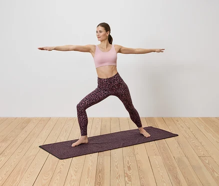 Frau in Sportbekleidung macht Yoga auf einer gemusterten Matte auf einem Holzboden vor einer weißen Wand.