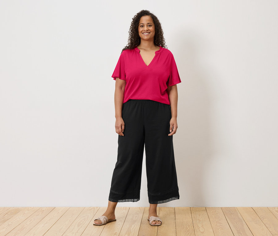 Frau mit lockigem Haar trägt ein pinkes T-Shirt, schwarze Culottes mit Spitzenbesatz und Sandalen vor einer weißen Wand.