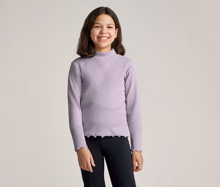 Lächelndes Mädchen mit braunen Haaren trägt ein lila Langarmshirt mit Rollkragen und schwarze Leggings vor einem hellen Hintergrund.