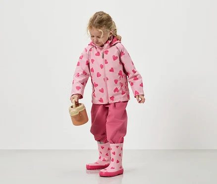 Ein kleines Mädchen schüttet Sand aus einem Spielzeugeimer, bekleidet mit einer rosa Jacke mit Herzmuster, einer passenden Hose und Gummistiefeln.