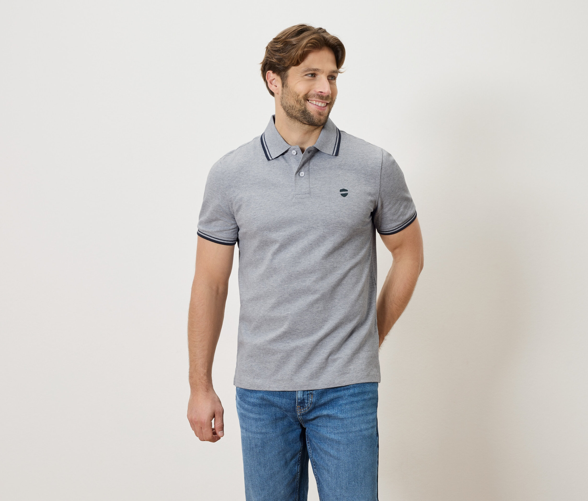 Mann mit grauem Poloshirt und Jeans, stehend vor einem neutralen Hintergrund.