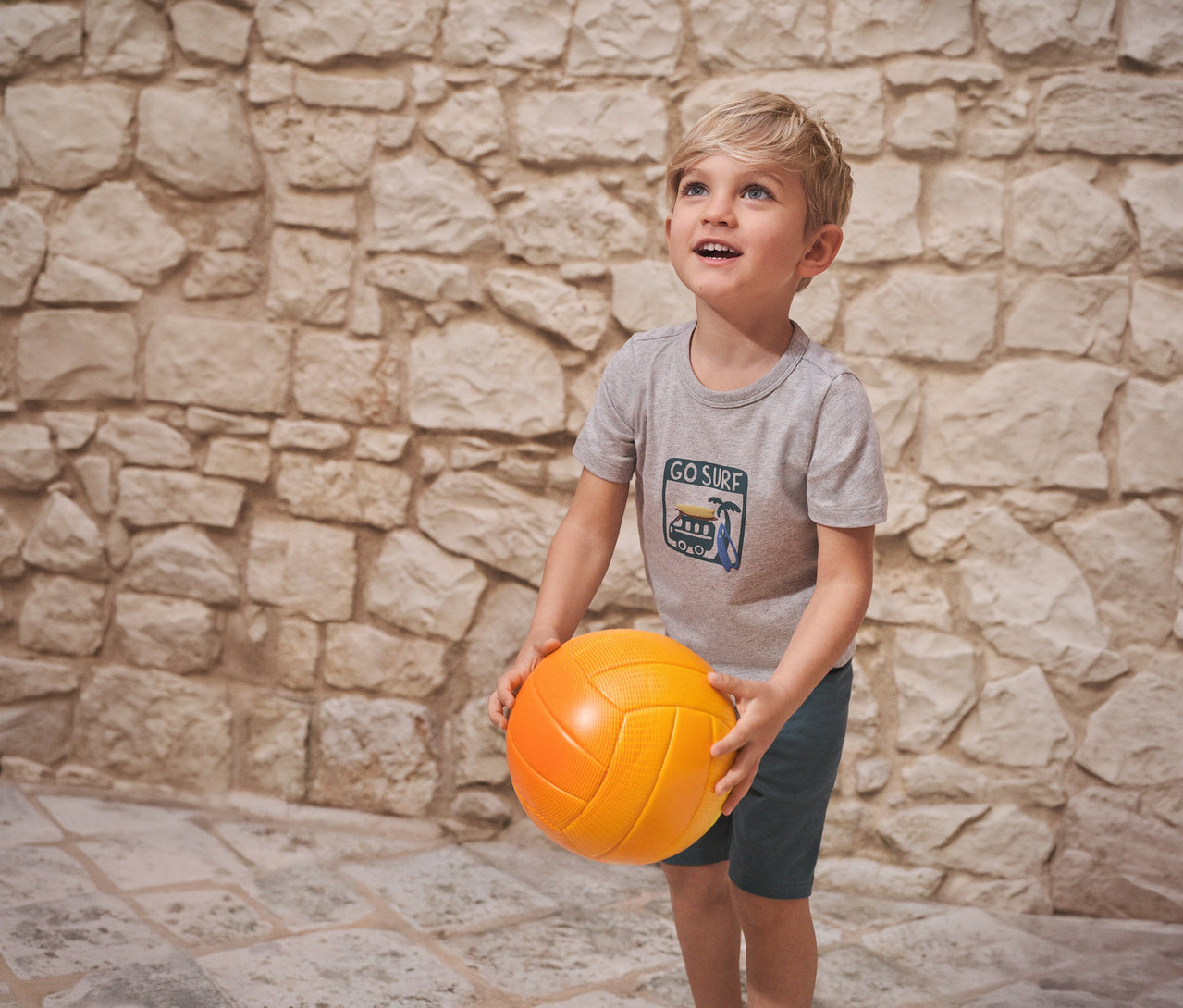 Junge hält einen orangefarbenen Ball und trägt einen kurzen Kinder-Pyjama von SCHIESSER vor einer Steinwand.