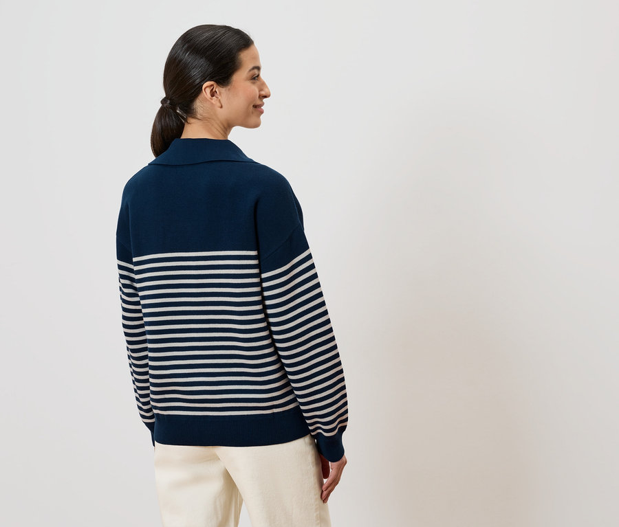 Frau von hinten, mit Pferdeschwanz, trägt einen dunkelblauen, gestreiften Pullover und helle Hose vor einem weißen Hintergrund.