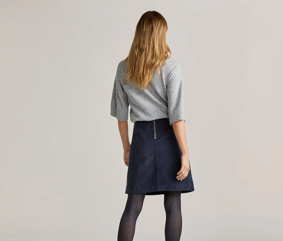 Rückansicht einer Frau mit blonden, schulterlangen Haaren, die einen grauen Pullover, einen marineblauen Rock und eine dunkle Strumpfhose trägt.
