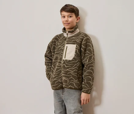 Ein junger Junge trägt eine olivgrüne Fleecejacke mit hellen Wellenmustern und hellen Jeans, steht vor einer hellen Wand. Die Jacke hat einen durchgehenden Reißverschluss und eine helle Brusttasche mit Reißverschluss.