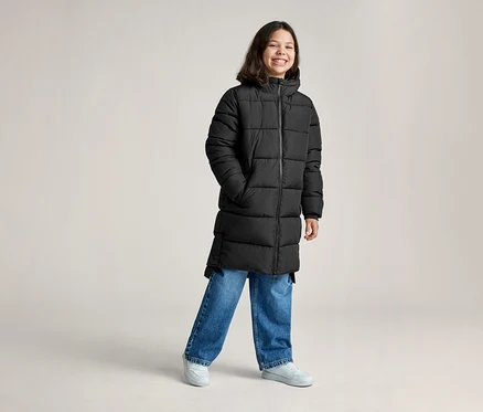 Ein lächelnder junger Mensch trägt einen langen schwarzen Steppmantel, blaue Jeans und hellgraue Sneaker und steht mit den Händen in den Taschen.