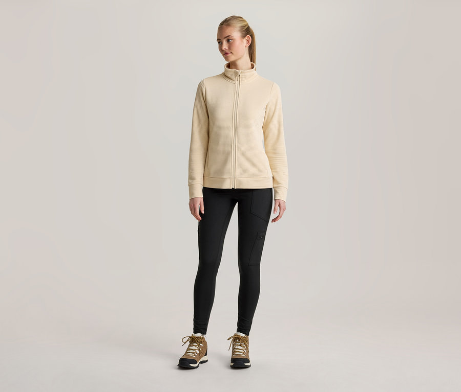 Eine junge Frau steht frontal, trägt eine hellgelbe Jacke mit durchgehendem Reißverschluss, schwarze Leggings mit Seitentaschen und braune Wanderstiefel mit weißen Schnürsenkeln. Ihr blondes Haar ist zu einem Pferdeschwanz gebunden.
