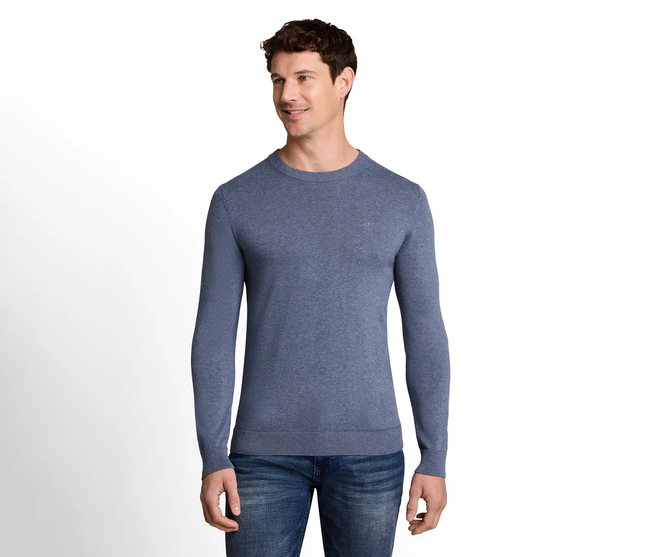Ein lächelnder Mann in einem blauen Langarmpullover und Jeans, der nach rechts blickt, steht vor weißem Hintergrund.