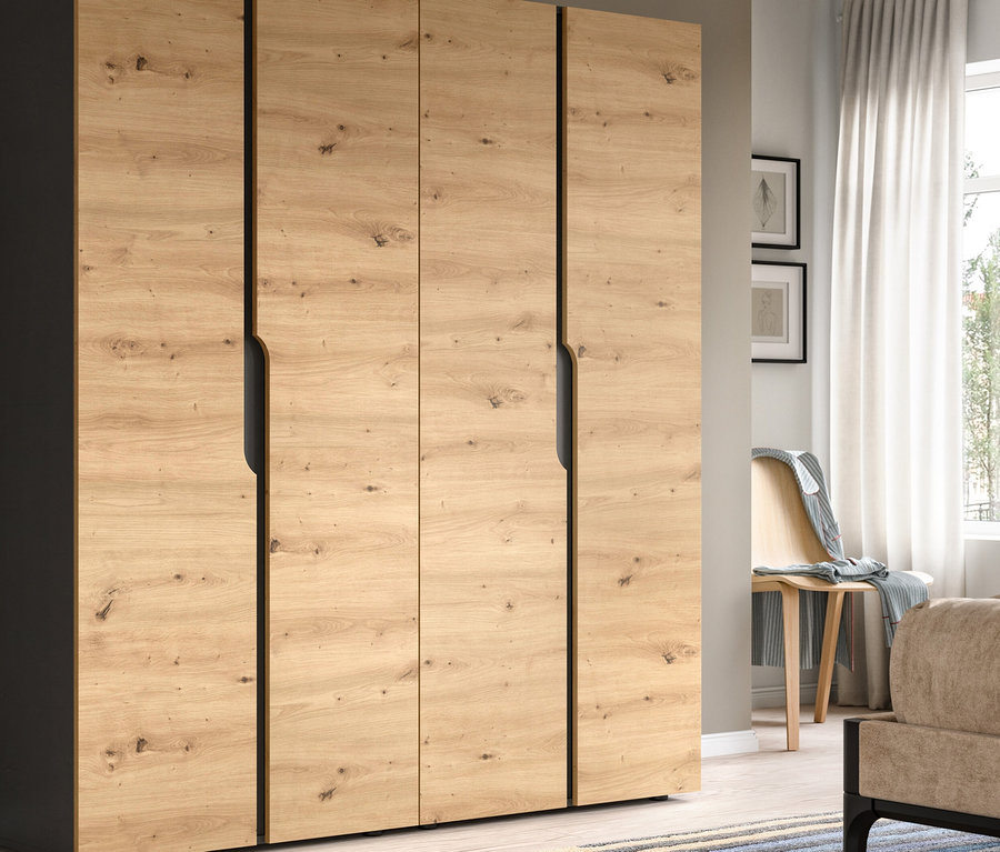 Ein großer viertüriger Kleiderschrank mit hellen Holzmaserungsfronten und dunklen Seitenpaneelen steht in einem Schlafzimmer. Rechts davon blickt ein Fenster mit weißen Vorhängen auf Grünflächen, daneben stehen ein Bett, ein Nachttisch mit einer weißen Lampe und ein Holzstuhl mit einer Decke.