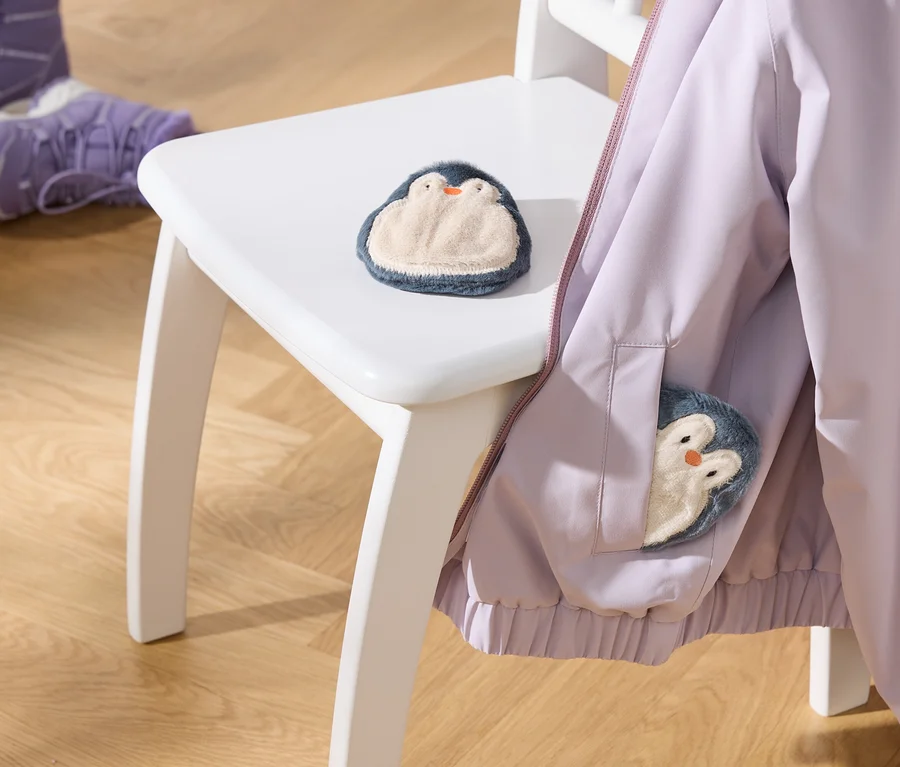 Eine hellviolette Jacke hängt auf einem weißen Stuhl, mit einem flauschigen Pinguin-Spielzeug auf der Sitzfläche und einem weiteren in der Jackentasche. Zwei lila Kinderstiefel stehen auf dem Holzboden.