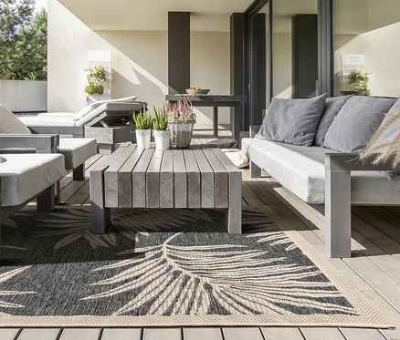 Graue Outdoor-Möbel auf einer Holzterrasse mit einem schwarz-beigen Teppich mit Blattmuster, Pflanzen und einem Esstisch im Hintergrund.