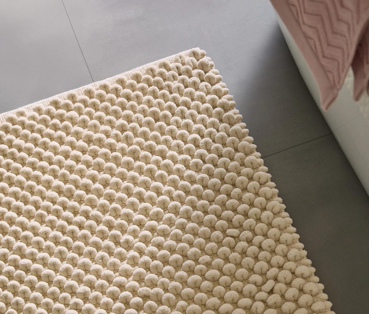 Nahaufnahme einer cremefarbenen Noppen-Badematte auf einem grauen Fliesenboden neben einer weißen Badewanne mit einem rosa Handtuch.