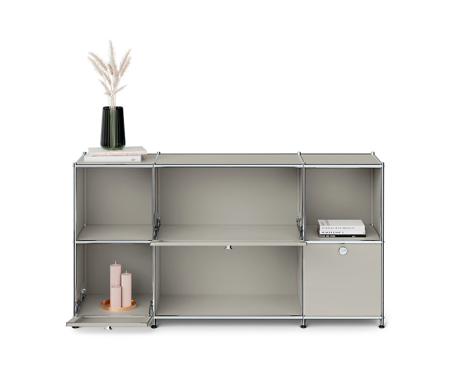 Graues modulares Regal mit mehreren Fächern. Oben links stehen eine Vase mit Pampasgras und Bücher, unten links sind drei rosa Kerzen auf einem Tablett sichtbar. Rechts ist ein geschlossenes Fach.
