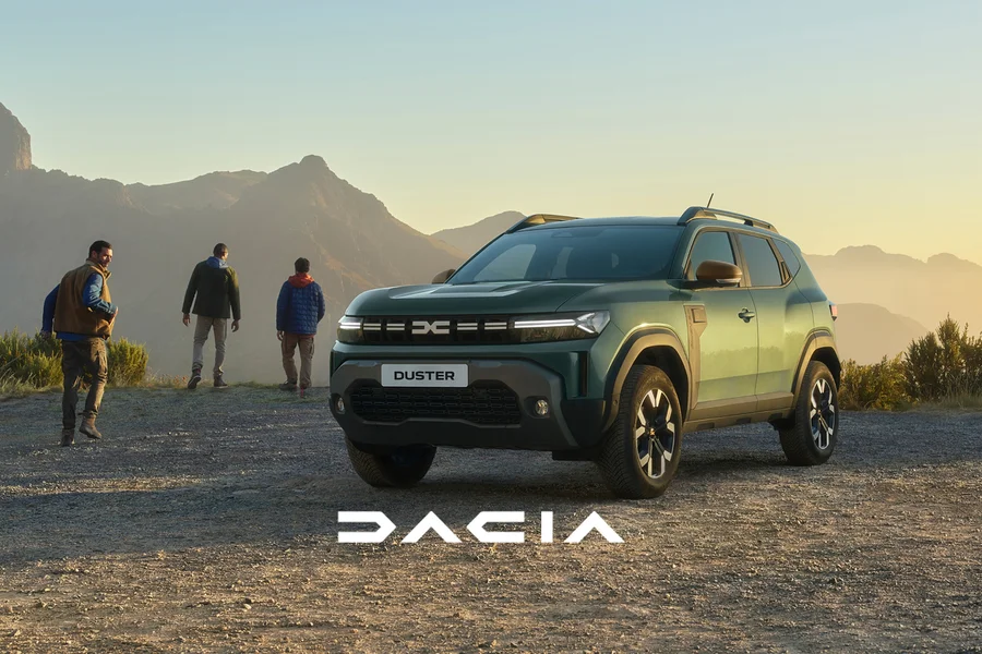 Ein grüner Dacia Duster SUV steht auf einem Schotterweg. Drei Männer stehen dahinter und blicken auf eine weite Berglandschaft im warmen Licht des Sonnenuntergangs. Unten ist das Dacia Logo zu sehen.