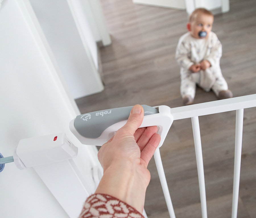 Eine Hand öffnet das weiße roba easySafe+ Tür-und-Treppen-Schutzgitter, während ein Baby mit Schnuller im Hintergrund auf dem Boden sitzt.
