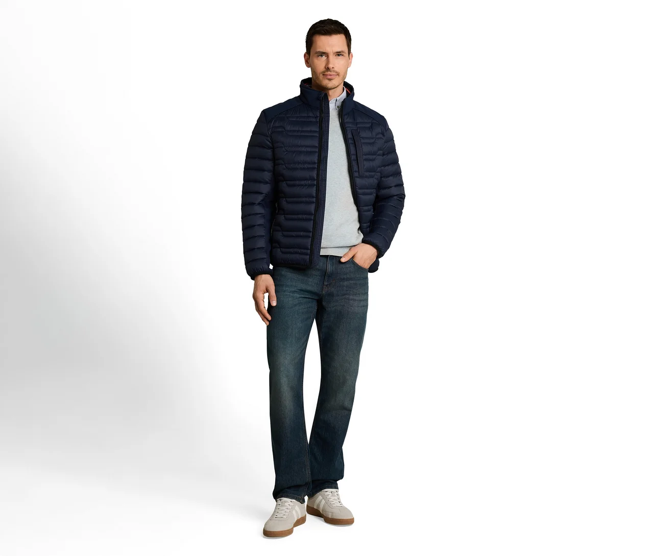 Mann steht in dunkelblauer Steppjacke, hellgrauem Langarmshirt, blauer Jeans und hellen Sneakern. Eine Hand steckt in der Hosentasche.