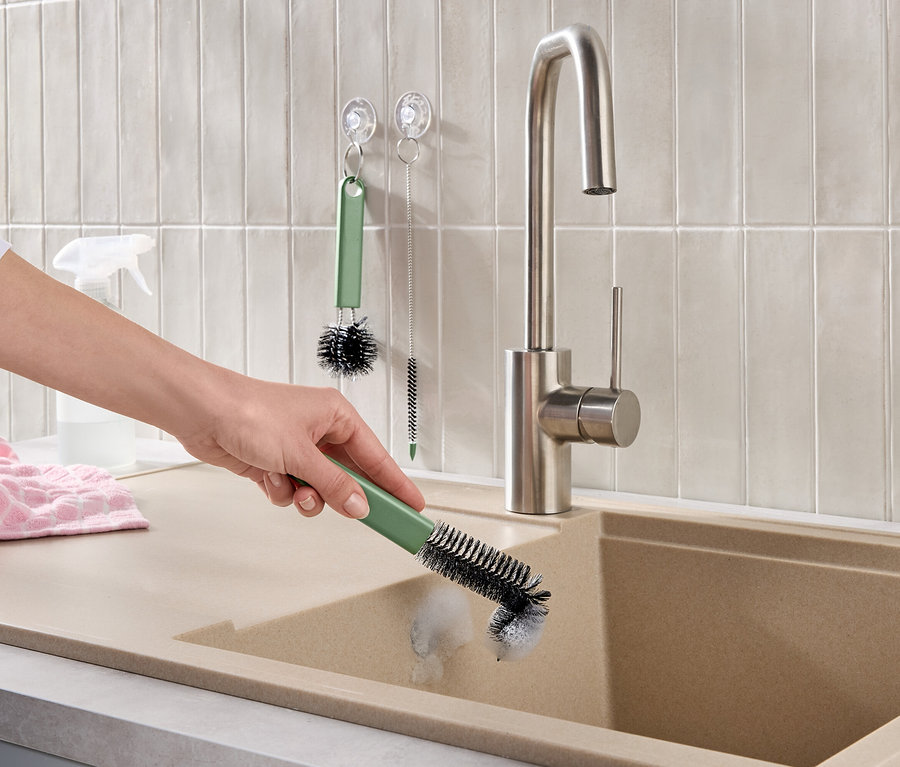 Hand reinigt beiges Spülbecken mit grüner Bürste. Ein Edelstahl-Wasserhahn, helle Wandfliesen, ein rosa Handtuch und weitere Reinigungsbürsten sind sichtbar.