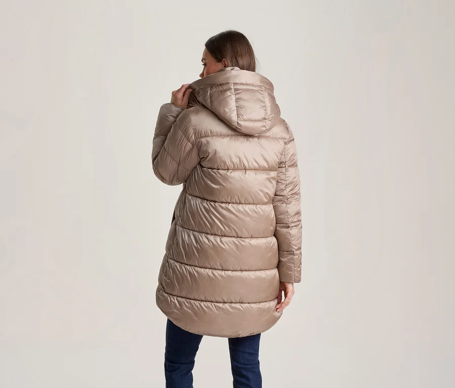 Frau von hinten, trägt eine hellbraune/beige Kapuzen-Steppjacke und dunkle Jeans. Sie hält den Kragen der Jacke mit der rechten Hand.