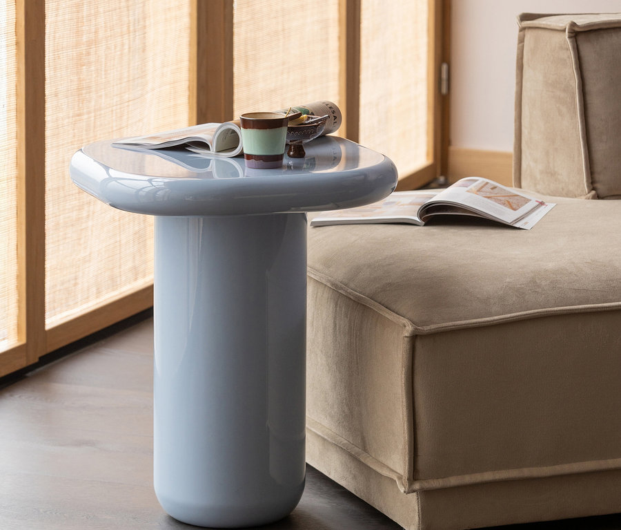 Ein hellblauer, glänzender zylindrischer Beistelltisch mit abgerundeter Platte, auf dem eine offene Zeitschrift, eine gestreifte Tasse und eine kleine Snackschale stehen, neben einem beigen modularen Sofa vor einem Fenster mit geflochtenem Rollo.