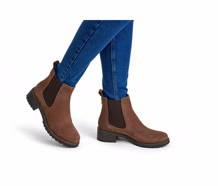 Eine Person trägt blaue Jeans und braune Wildleder-Chelsea-Boots mit elastischen Seiteneinsätzen und einer klobigen Sohle auf weißem Hintergrund.