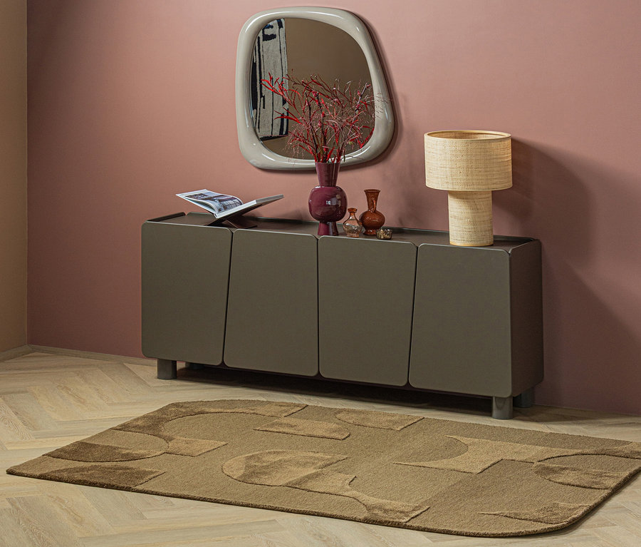 Ein brauner WOOOD Teppich »Livo« mit abstrakten Mustern liegt auf hellem Holzboden vor einem dunkelgrauen Sideboard. Darauf stehen eine offene Zeitschrift, eine lila Vase mit roten Zweigen, kleine braune Vasen und eine geflochtene Tischlampe. Ein unregelmäßig geformter Spiegel hängt an der rosa Wand.