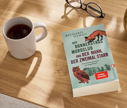 Auf einem Tisch liegen ein Buch, eine Tasse mit einem Getränk, eine Brille und eine Vase.