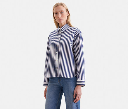 Blonde Frau in einem blau-weiß gestreiften Langarmhemd und blauen Jeans, blickt nach vorne.