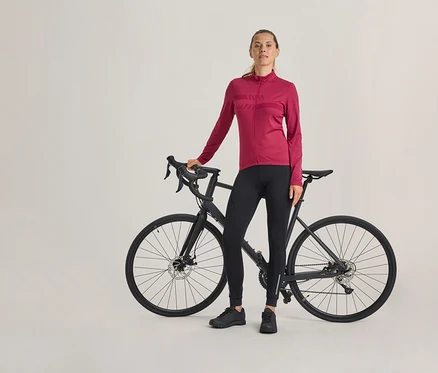 Frau steht neben einem schwarzen Rennrad, trägt ein magentafarbenes langärmeliges Radtrikot, schwarze Radhose und schwarze Schuhe.
