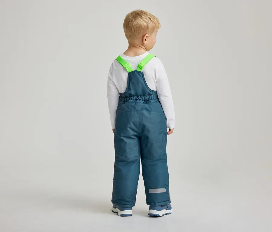 Ein kleines Kind mit blondem Haar steht mit dem Rücken zur Kamera und trägt ein weißes Langarmshirt und blaugrüne Schneehosen mit leuchtend grünen Hosenträgern. Die Schneehose hat einen Gummizug an der Taille und reflektierende Streifen an den Knöcheln.