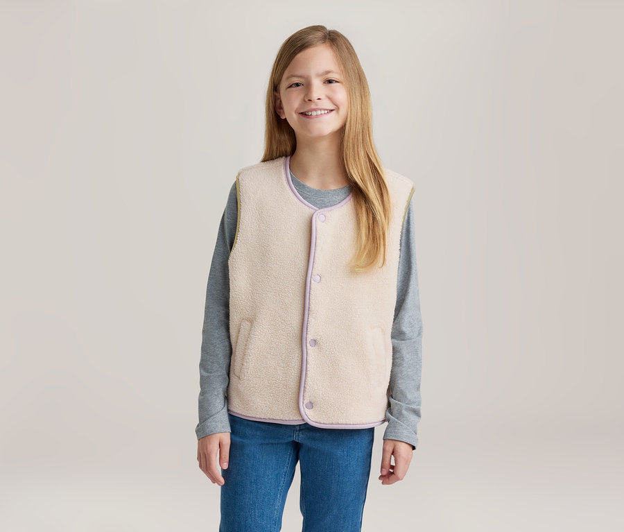 Junges Mädchen mit langen blonden Haaren lächelt und trägt eine cremefarbene Fleeceweste mit lila Besatz über einem grauen Langarmshirt und blauen Jeans.