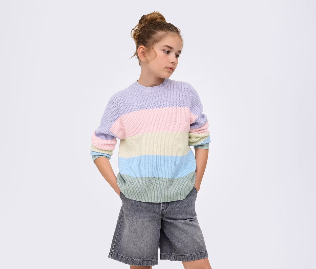Mädchen mit Dutt trägt einen gestreiften ONLY GIRLS Sandy Strickpullover in Lavendel, Rosa, Gelb, Hellblau und Hellgrün sowie graue Jeans-Shorts, Hände in den Taschen, blickt zur Seite vor weißem Hintergrund.