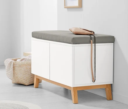 Eine weiße Sitzbank mit Stauraumfächern und grauem Kissen. Auf der Bank liegt eine beige Handtasche.
