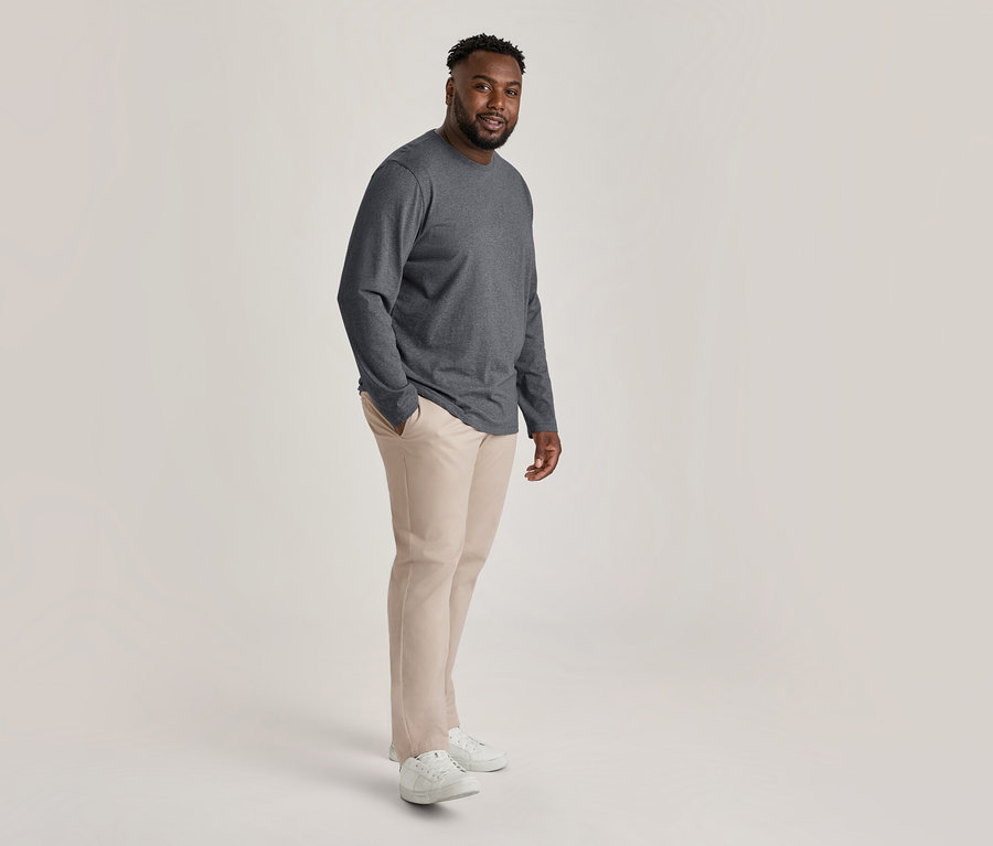 Lächelnder Mann trägt dunkelgraues Langarmshirt, beige Chino und weiße Sneaker, eine Hand in der Hosentasche.