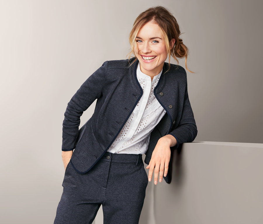Frau lächelt, an eine Wand gelehnt, trägt einen Sweatblazer und eine Hose.