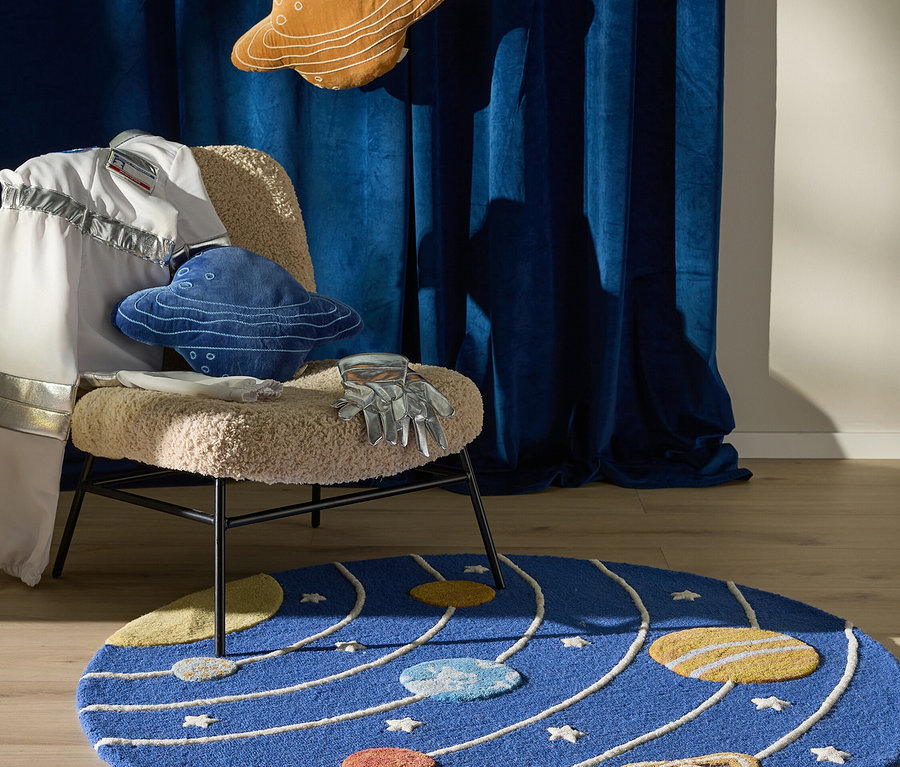 Ein Weltraum-Themenzimmer mit einem blauen runden Teppich mit Sonnensystem-Design auf einem Holzfußboden. Ein Stuhl mit flauschigem Sitz trägt einen weißen Astronautenanzug, ein blaues UFO-Kissen und silberne Handschuhe. Darüber hängen ein braunes Saturn-Kissen und ein Raketen-Kissen vor dunkelblauen Vorhängen.
