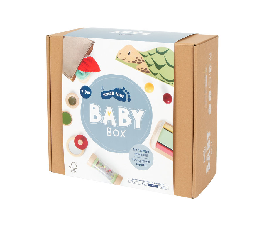 Ein brauner Karton für die „Small Foot Baby Box“ zeigt auf der Vorderseite Illustrationen verschiedener Babyspielzeuge. Das Etikett zeigt „BABY BOX“ mit einer Altersangabe „7-9m“, einem FSC-Logo und dem Text „Mit Experten entwickelt!“. Abgebildete Spielzeuge sind eine Holzschildkröte, Stoffartikel, bunte Bälle, sensorische Scheiben, ein Rasselrohr und Holzbausteine.