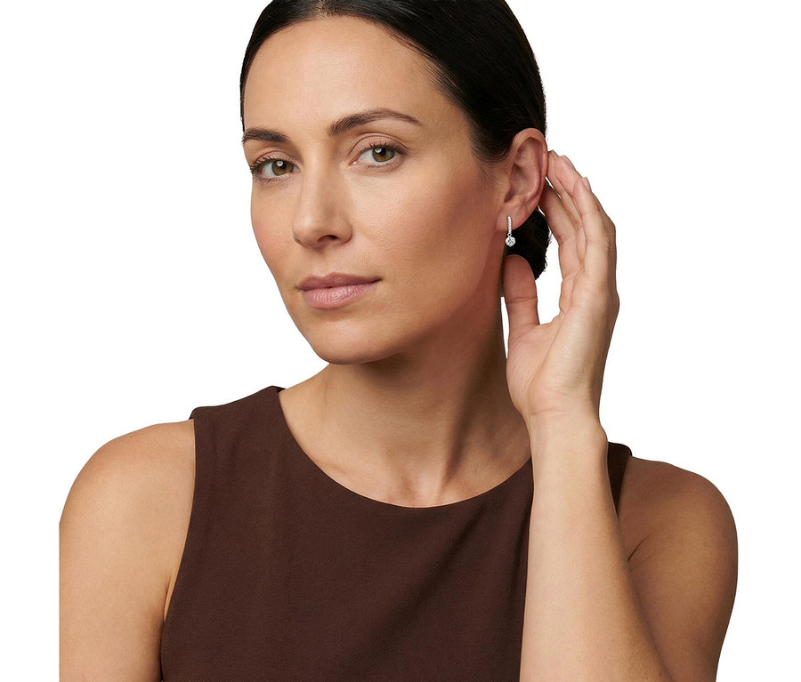 Eine Frau mit zurückgebundenem dunklem Haar und einem braunen ärmellosen Oberteil blickt in die Kamera. Sie trägt einen Amor Ohrschmuck am rechten Ohr und berührt ihr Ohr mit der rechten Hand.