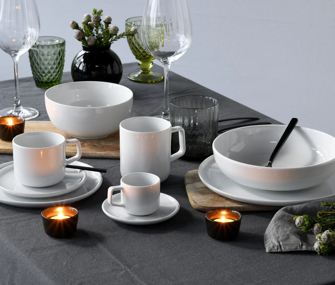 Gedeckter Tisch mit weissem, geripptem creaTable Service »Session White«, bestehend aus Schüsseln, Tassen und Bechern, auf dunkelgrauer Tischdecke. Dazu klare Weingläser, grüne und graue Trinkgläser, schwarze Teelichter und eine Vase mit Pflanzen.