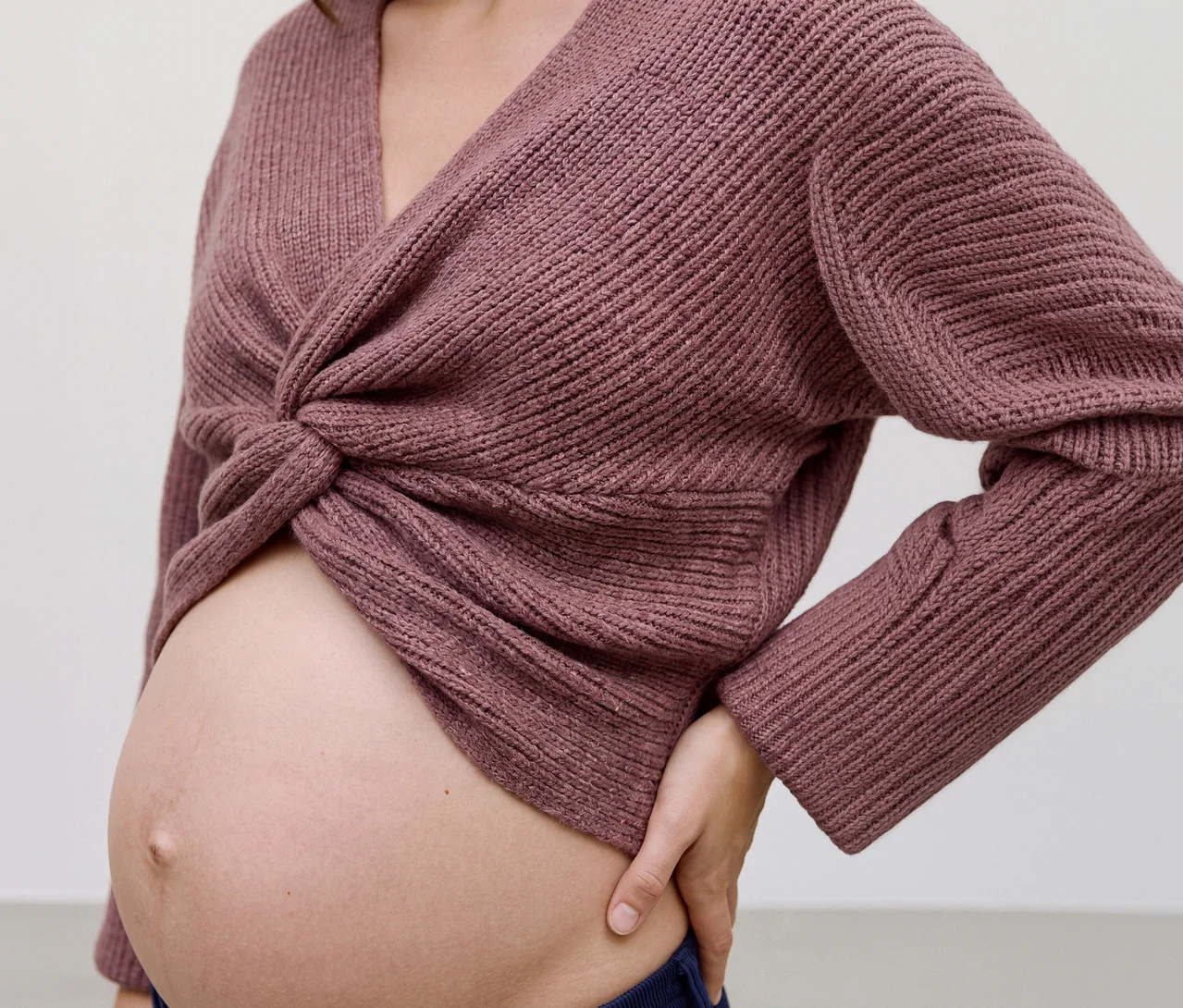 Eine schwangere Frau trägt ein mauvefarbenes MAMALICIOUS Cropped Stricktop »SVALA« mit gedrehter Vorderseite, das ihren Bauch freilegt. Ihre linke Hand ruht auf ihrer Hüfte.