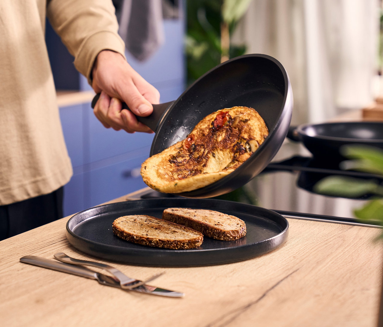 Eine Person schiebt ein Omelett aus einer schwarzen Henckels by ZWILLING »Breeze« Bratpfanne auf einen Teller mit zwei Scheiben Toastbrot. Besteck liegt auf der Holztheke.