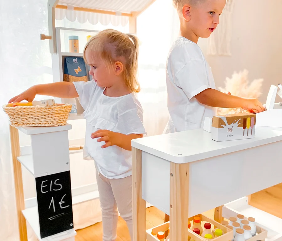 Zwei Kinder spielen mit einem small foot Kaufladen kompakt »fresh«. Ein Mädchen greift in einen Korb. Ein Junge steht an der Theke.