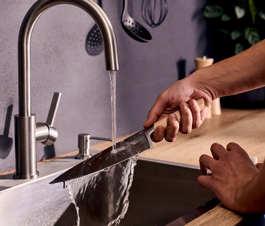 Eine Hand hält ein Henckels by ZWILLING Pakka Messer mit Holzgriff unter fließendem Wasser aus einem Küchenhahn und spült die Klinge in einem Edelstahlwaschbecken ab.