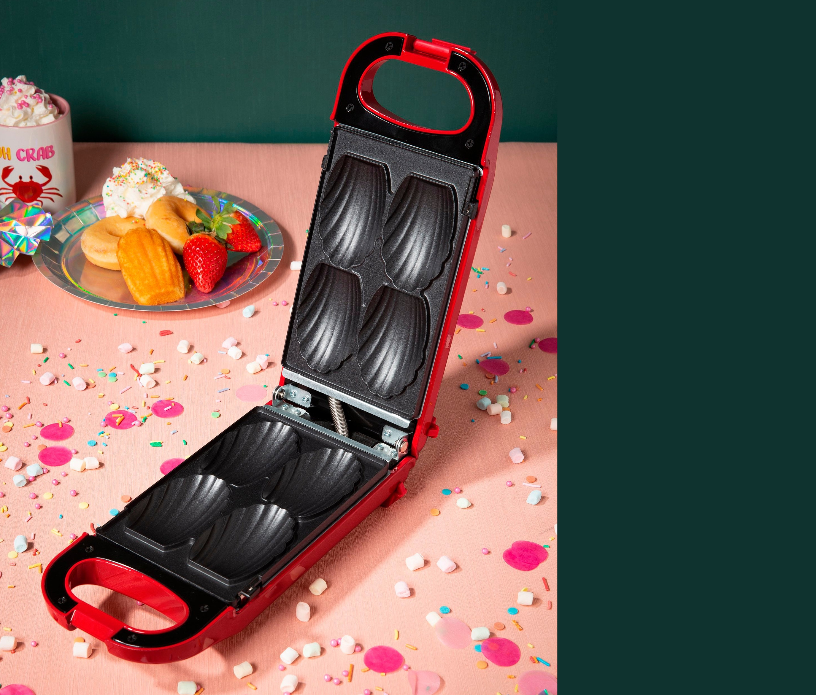 Ein offener, roter 6-in-1-Snackmaker auf rosafarbenem Hintergrund mit Konfetti, Teller mit Donuts, Erdbeeren und Madeleines.