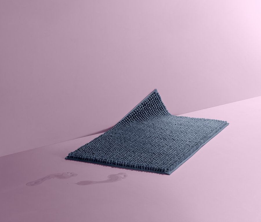 Eine blaue Badematte, ca. 50 x 80 cm, liegt auf einem rosafarbenen Untergrund. Davor sind nasse Fußspuren zu sehen.