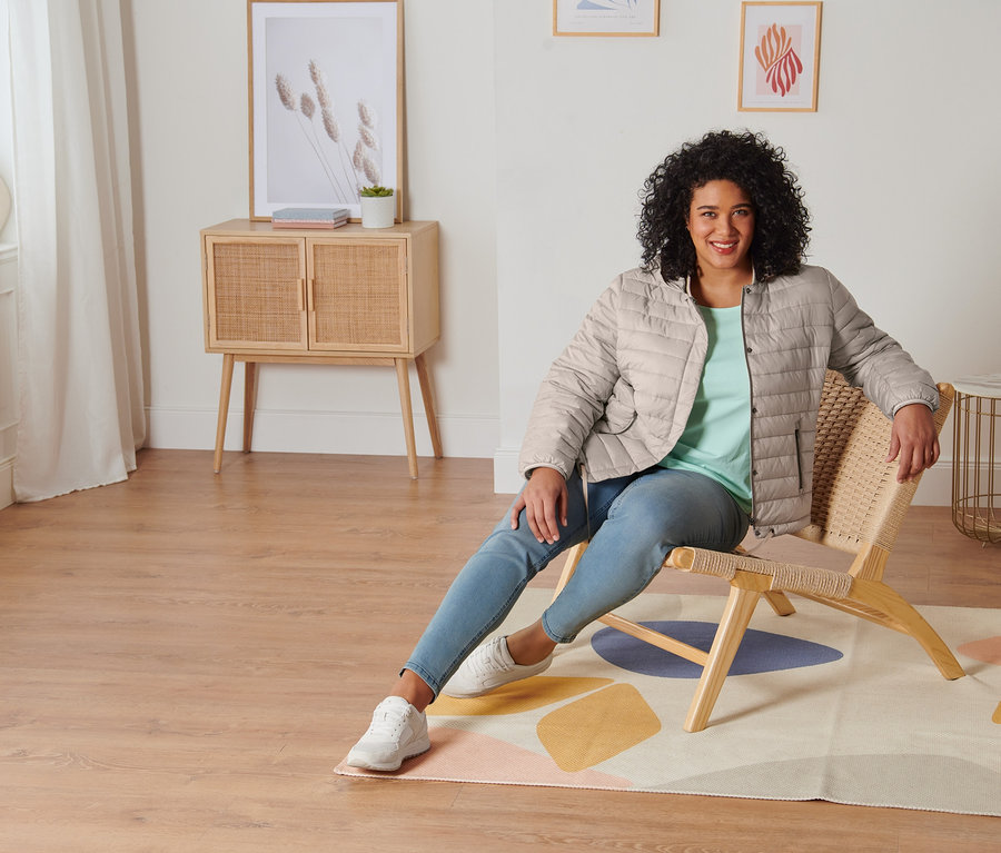 Frau sitzt auf einem Stuhl und trägt ein mintfarbenes Shirt mit 3/4-Arm, Treggings, Veloursleder-Sneaker und eine Steppjacke mit Kapuze.
