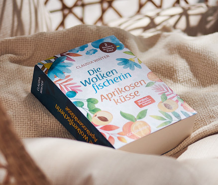Doppelroman »Die Wolkenfischerin« und »Aprikosenküsse« liegt auf einem beige-farbenen Kissen in einem Rattansessel.